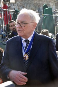 A photograph of Warren Buffet, dressed in a blue suit jacket, white shirt, and dotted tie, with his hand in front of his torso. His Presidential Medal of Freedom is around his neck. Behind him a grey brick building and a two people are visible.