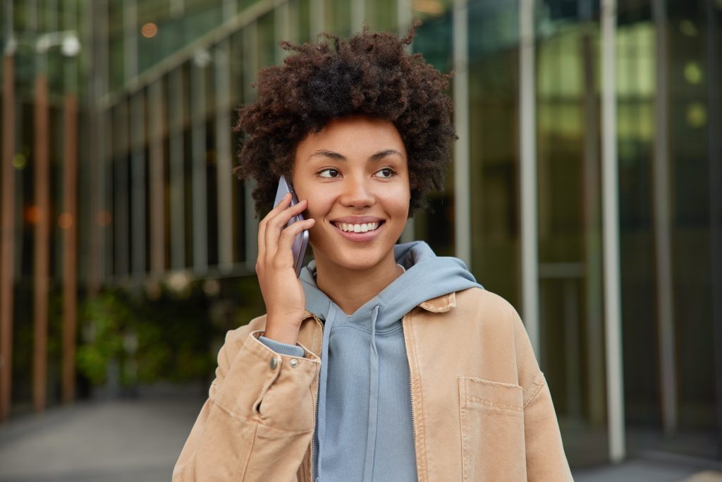 Modern technologies and people concept. Happy young curly haired woman enjoys cellular communication smiles positively dressed casually poses outdoors against blurred background makes telephone call