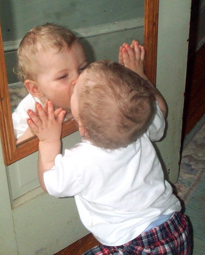 A blonde baby in a white t-shirt kisses themselves in a mirror.