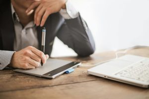 A focused individual writing on a tablet while a laptop sits in front of them.