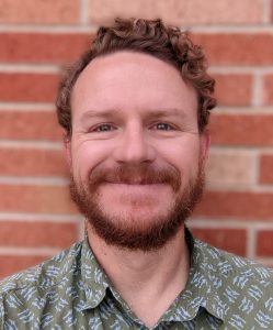 Picture of Eric Atkins wearing a green collared shirt and standing in front of a brick wall.