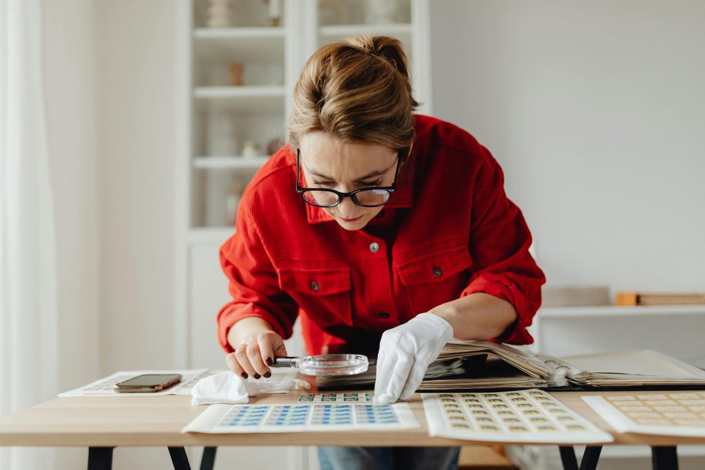 Woman in red shirt leans over a collection of small objects and examines them with a magnifying glass.