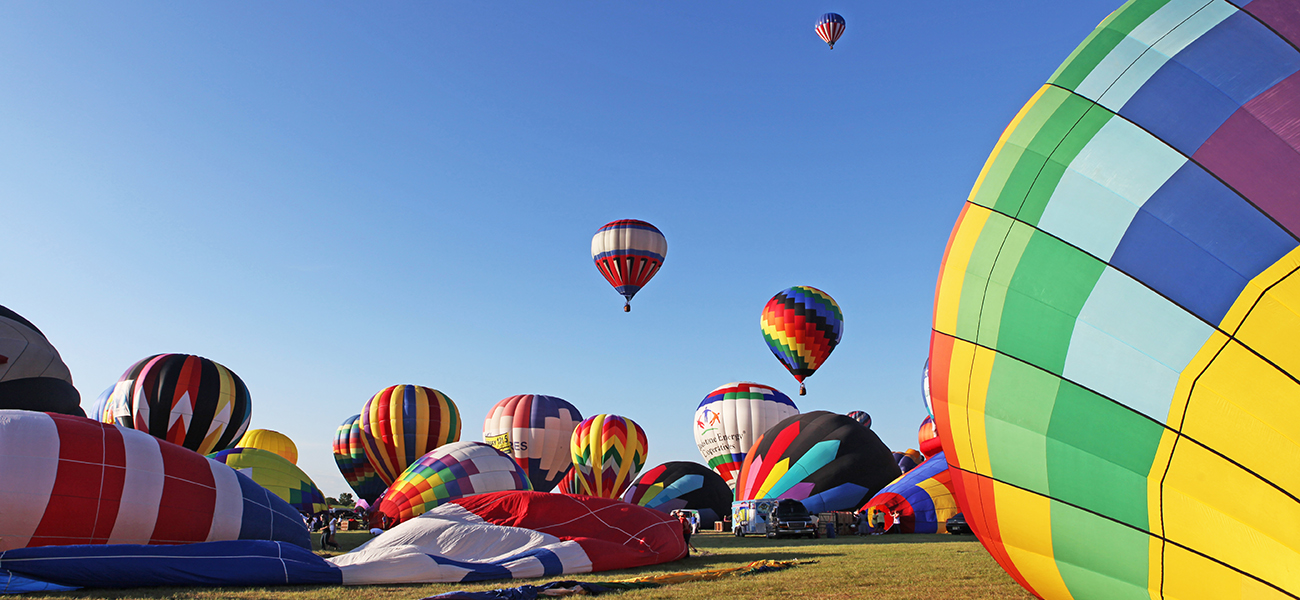 A photograph shows about twenty colorful hot air balloons at varying stages of inflation. Some are deflated, while others are inflated. Three of the balloons are off the ground and are visible against a bright blue sky.