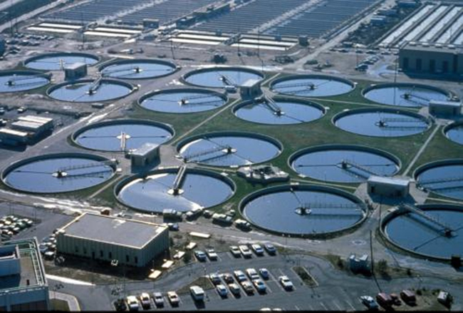 A color photograph is shown of a high volume wastewater treatment facility. Nineteen large circular pools of water undergoing treatment are visible across the center of the photograph. A building and parking lot are visible in the foreground.