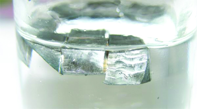 A glass container that is half filled with a colorless liquid is shown. Blocks of a shiny silver solid float on top of the liquid in the container.