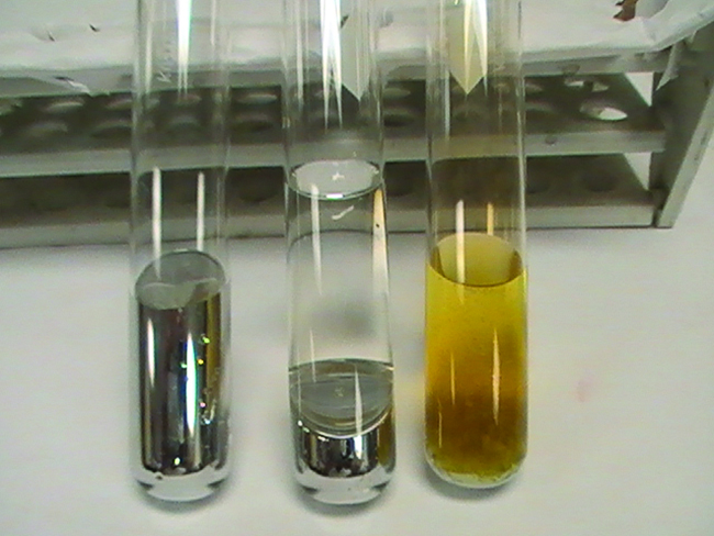 Three test tubes are shown in a photo. The left tube contains a metallic liquid. The middle tube contains a metallic liquid under a layer of clear, colorless liquid. The third tube contains a whitish solid under a layer of yellowish liquid.