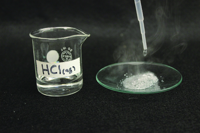A photo shows a beaker that contains a clear, colorless liquid. It is labeled, “H C l ( a q ).” Beside the beaker is a watch glass with a dropper above it. The dropper is releasing liquid into a fizzing liquid. The fizzing liquid is releasing a white gas.