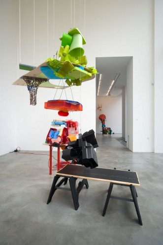 Bare room with a black table in the center and hangings of green plastic above, with a red table covered in miscellaneous items in the background