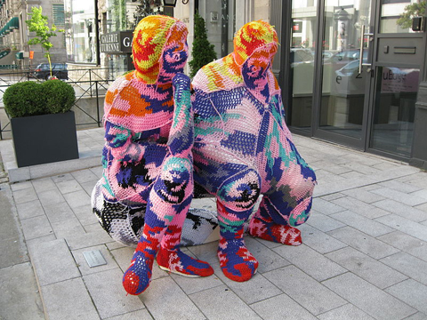 Two seated human figures covered in crocheted blankets of yellow, blue, pink, orange, red