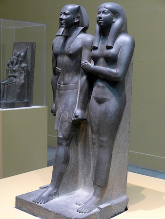 Stone statue of Egyptian king with queen holding his arm and waist