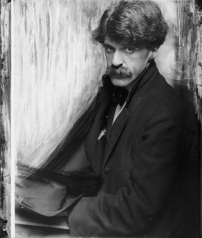 Man with a thick mustache, sitting before a white curtain and holding a hat