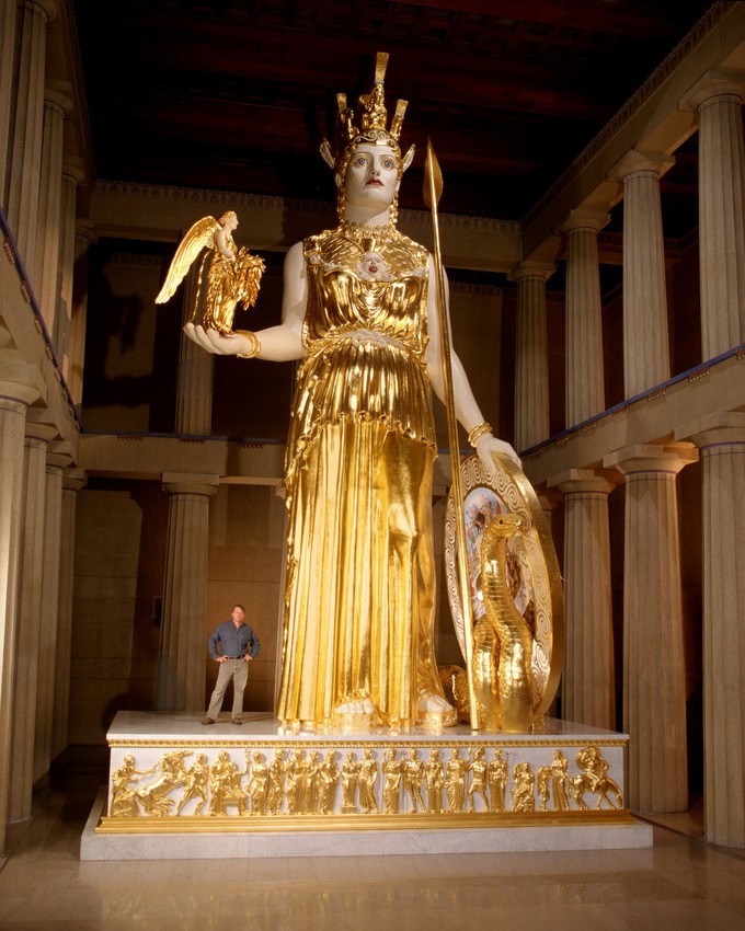 Tall statue of a woman with a golden armored dress, shield, and spear, with an angel in one hand