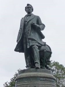 Dark gray statue of a man in a coat stepping forward against a gray sky