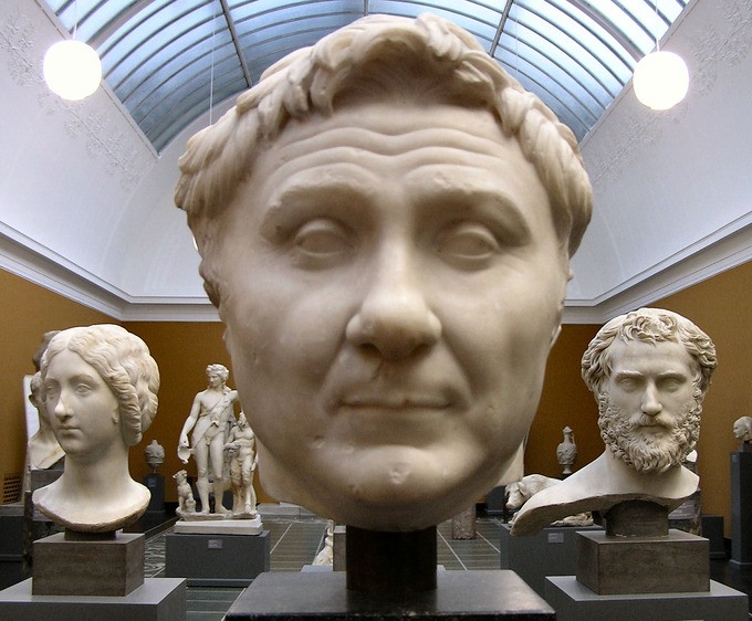 Large head on a pedestal in a room of other heads and statue figures