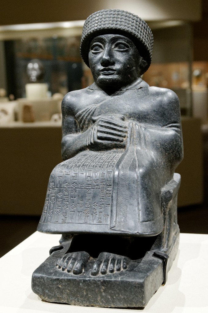 Stone man seated with hands folded and wearing a hat