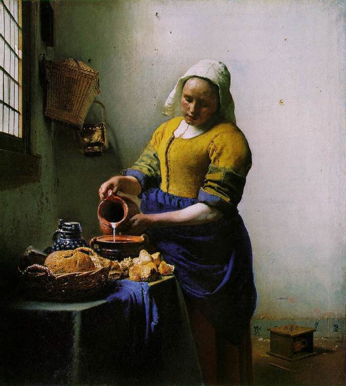Woman in a handkerchief and a yellow and blue dress pouring milk in a bowl beside stacks of bread