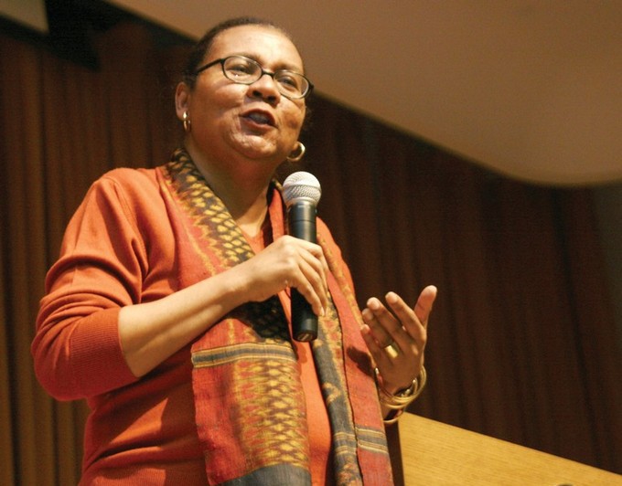 Woman wearing glasses, gold jewelry, and a long scarf speaking into a microphone