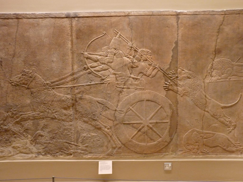 Carving in brown stone of soldiers in a chariot shooting arrows and staving off a lion with spears