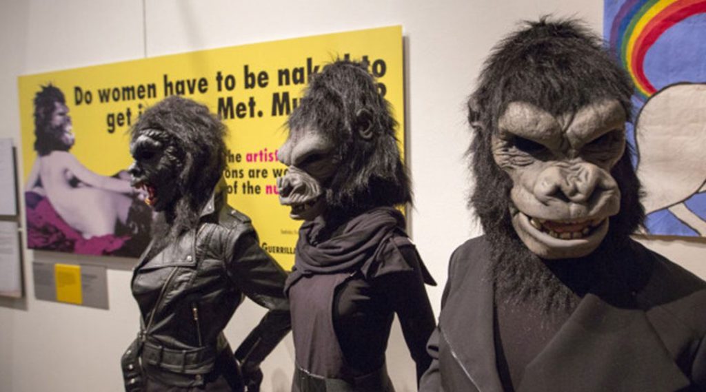 Three human sized gorillas in black outfits in front of a yellow sign on the wall