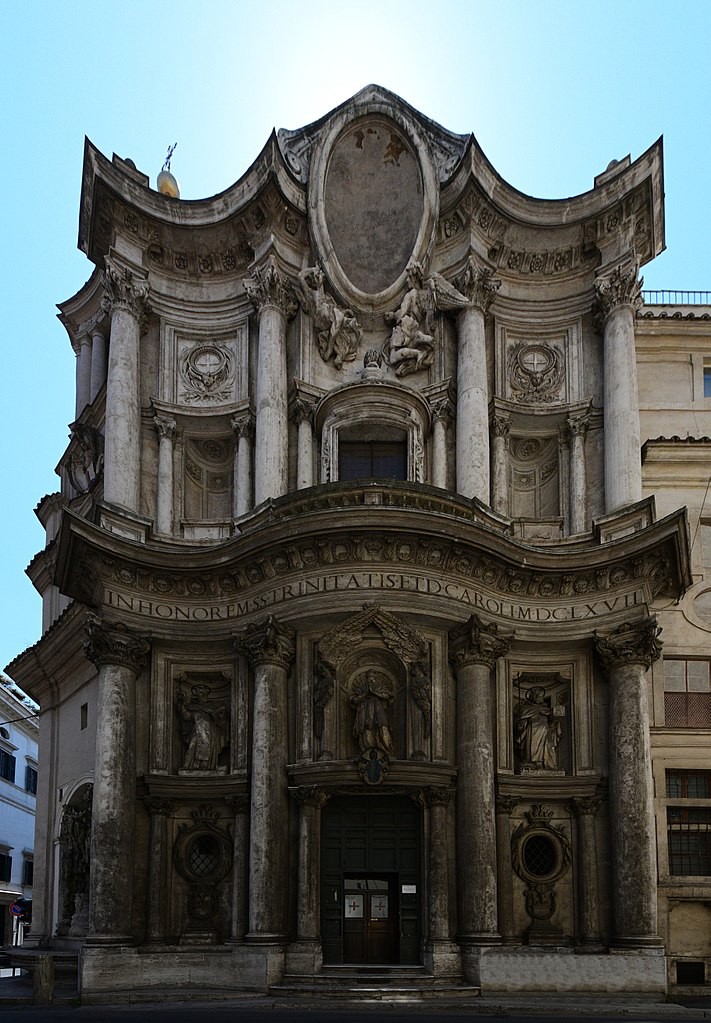 Curved dark stone building with statues and Latin writing on the front among several pillars