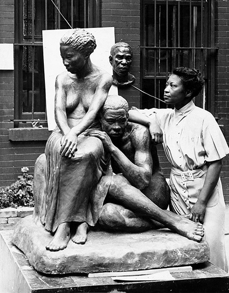Dark woman in a dress leaning against a statue of two dark-skinned naked people