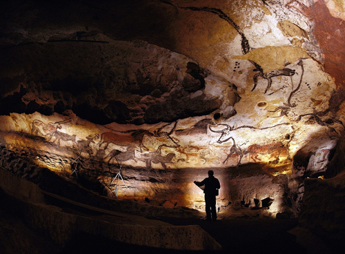 Man standing with a map looking at a cave wall covered in paintings of animals