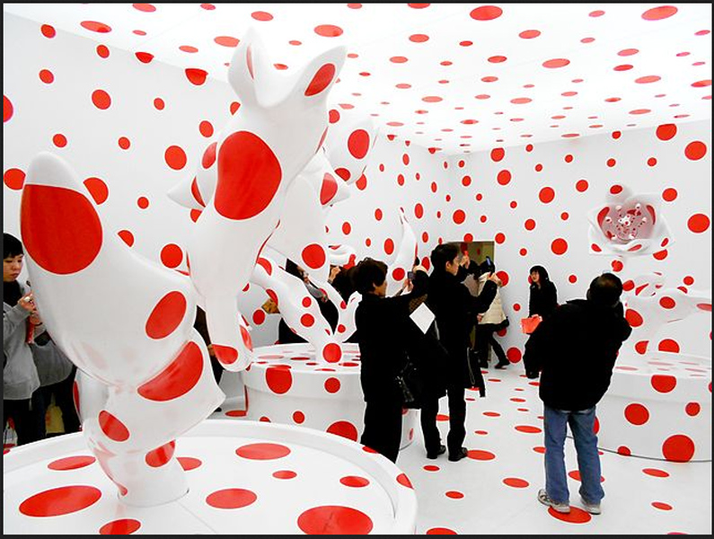 People in a white room covered with red spots including statues of the same