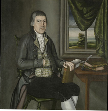 Elderly man seated at a desk with his hand in his vest and a book held on his lap