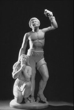 Stone statue of a man in shorts holding up a fist with a broken shackle and laying his hand on the shoulder of a kneeling woman praying