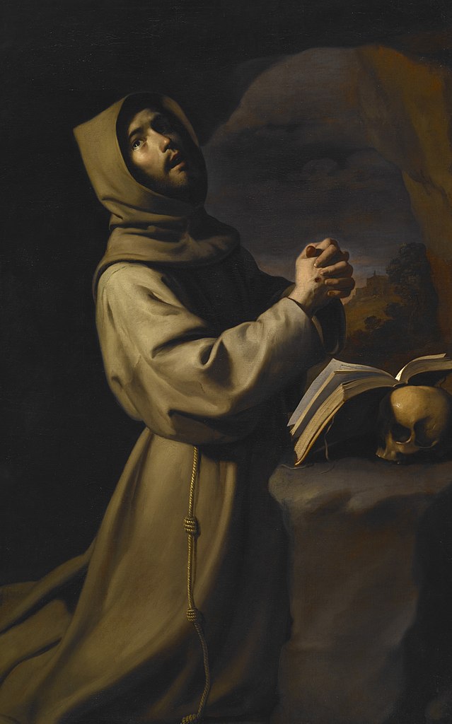 Man in a robe and hood kneeling before a book with hands clasped and looking upward