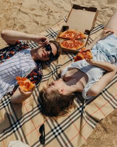 Two people laying down on a picnic blanket holding pizza