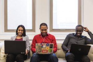 Three young professionals are sitting on a couch working on their laptop computers. They are smiling as they work.