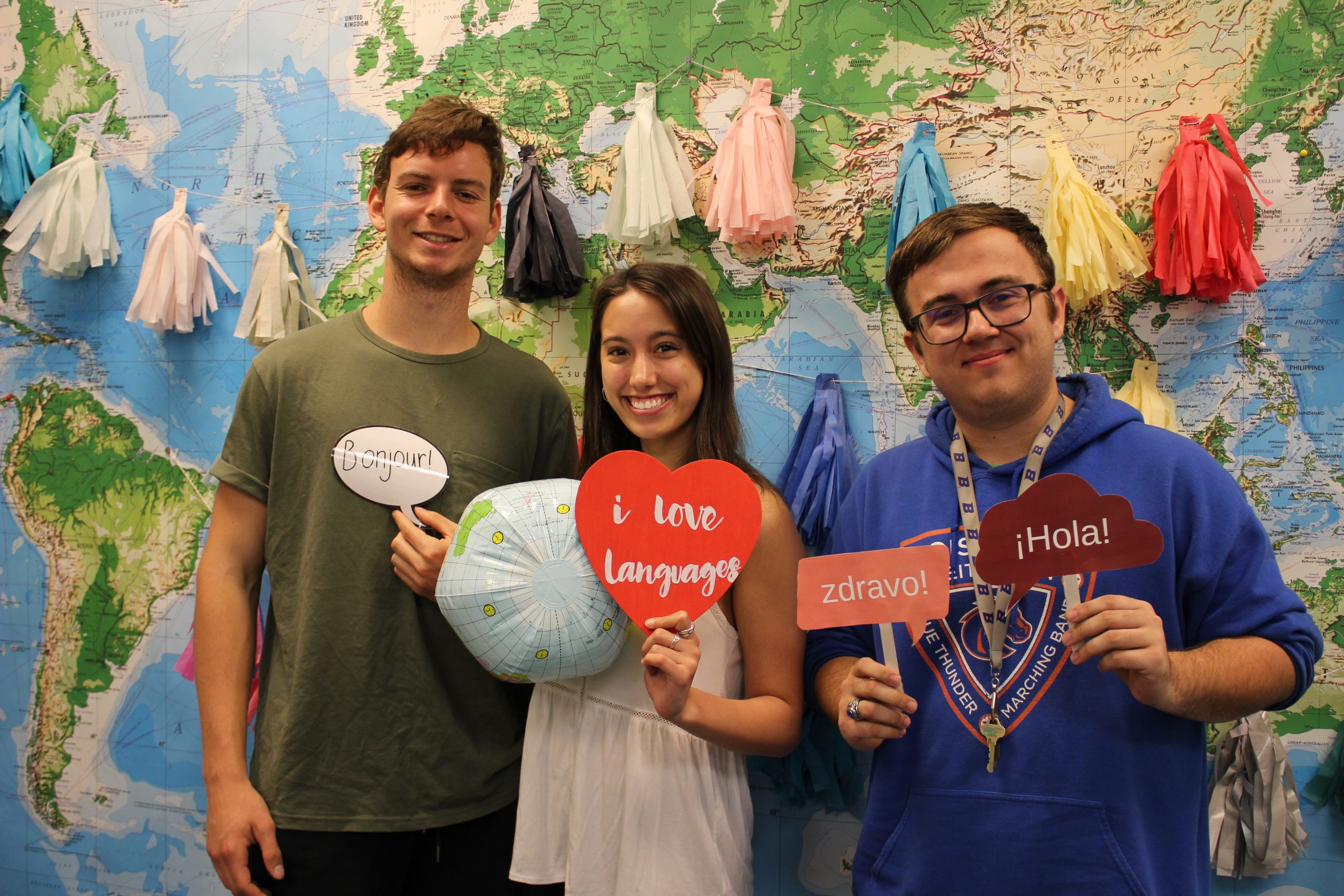 Photo of three language students holding props in front of a map