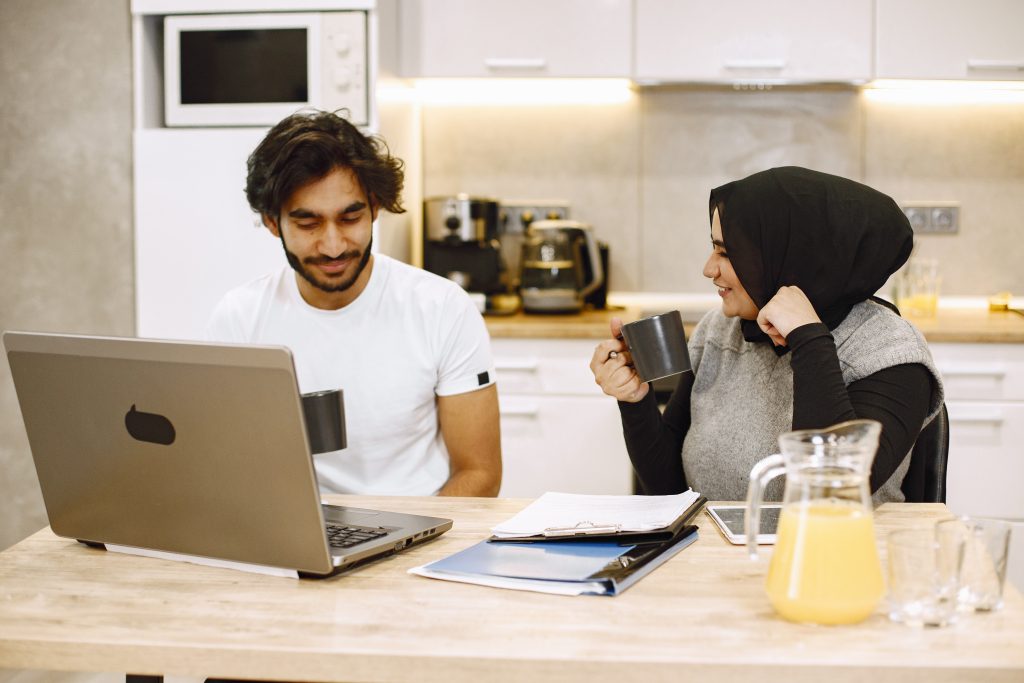Beautiful young couple using a laptop, writing in a notebook, sitting in a kitchen at home. Arab girl wearing black hidjab.