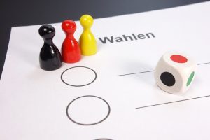 Photo of three game pieces (black, red, yellow), a colored dice and a paper with Wahlen (elections) written on it.(vote) on it.