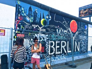 Photo of two women standing in front of a spray painted wall with the words Berlin and New York.
